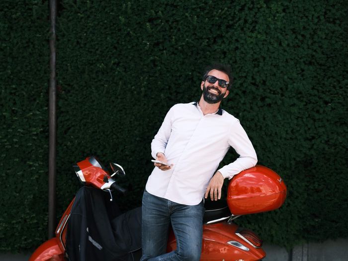 Simon Zaoui, adossé à un scooter rouge devant une haie, chemise blanche, lunettes de soleil, éclat de rire
