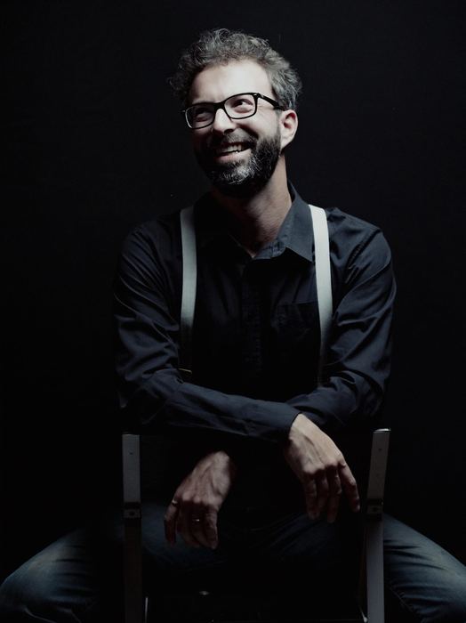 Simon Zaoui, assis sur une chaise, bretelles, grand sourire, regard levé